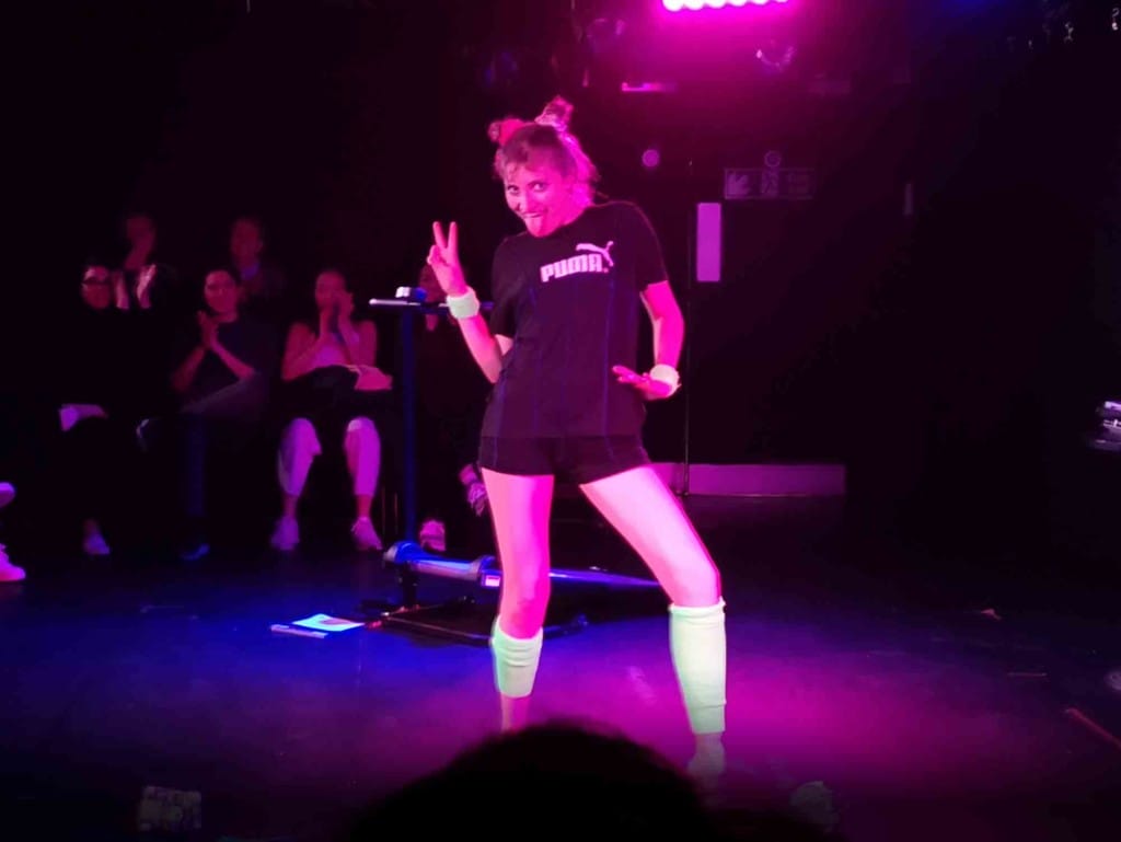 Dressed in gyn-wear, with neon green sweatbands and leg warmers, performer Frankie Thompson looks at the camera, posing and giving a victory sign. 