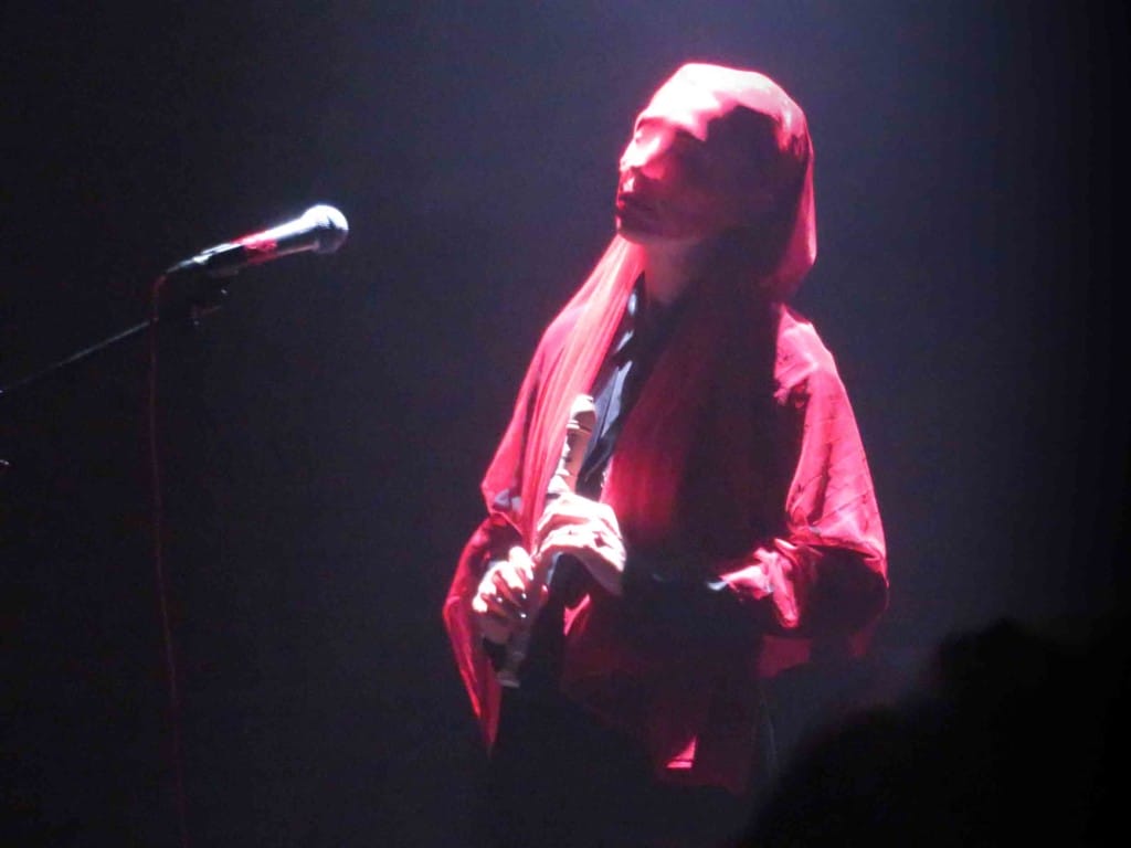 In a spotlight against a black background we wee a human figure holding a recorder. They are dressed in red with red gauze draped across their face.