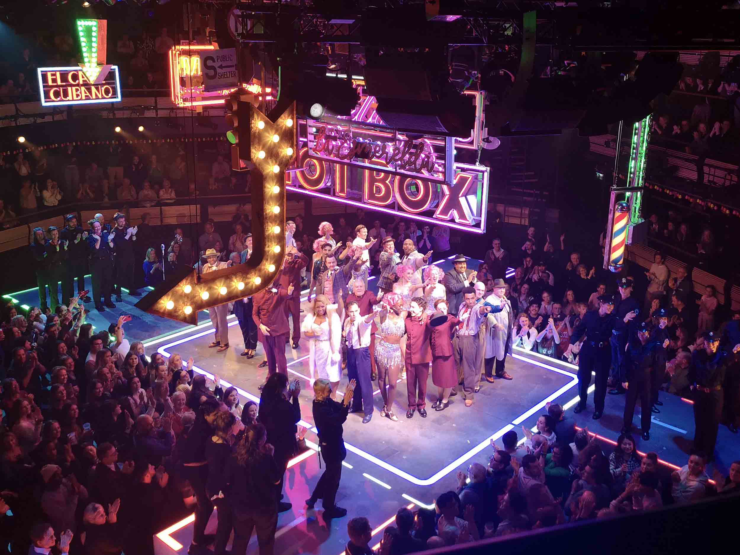 Looking down on the large cast of Guys and Dolls, closely huddled into a sqaure on the stage, all with one arm aloft. To all sides we see crowds of clapping people. Above them all are numerous neon signs. 