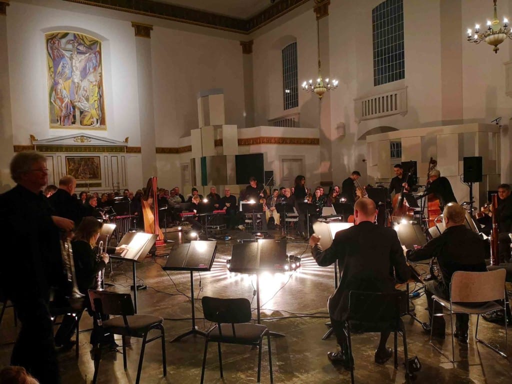 Interior of a white walled church. Musicians sit on chairs in a circle around a floor level fan of white light. On the wall is a painting of a crucified Christ.