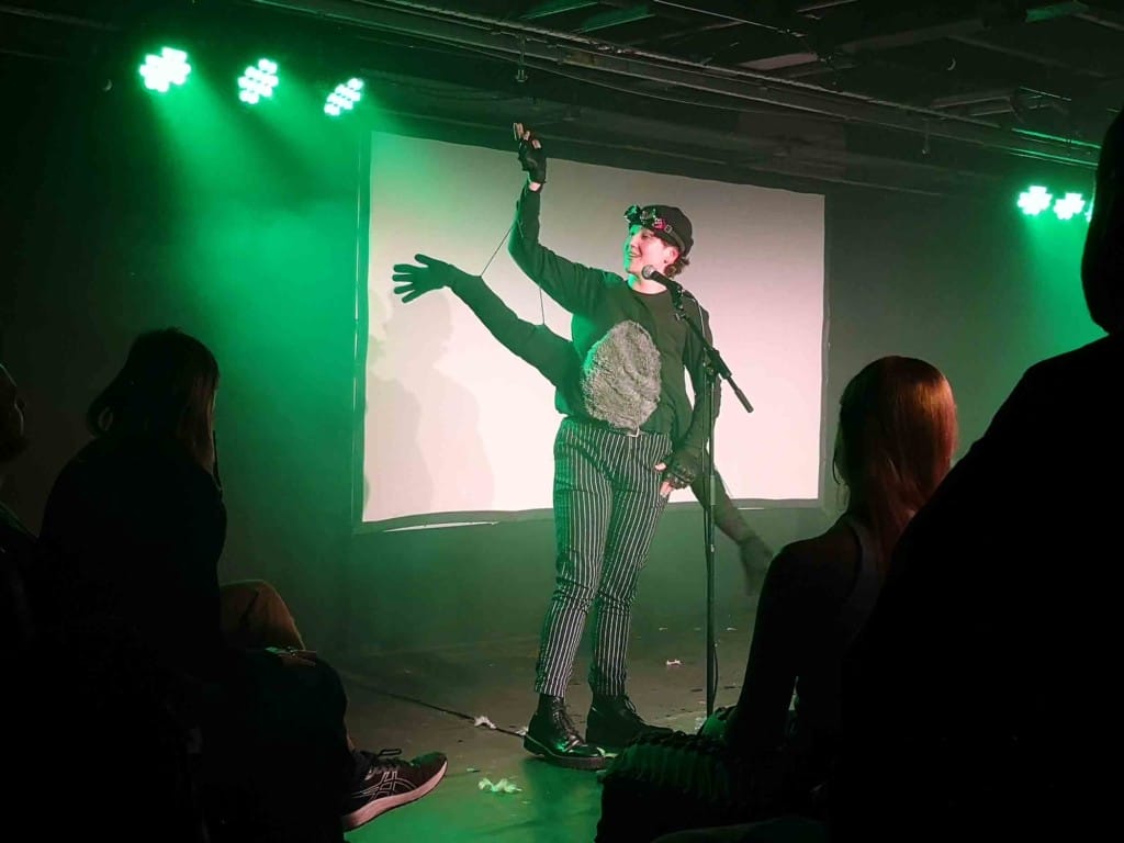 In a small venue, lit by green spotlights, performer Liv Ello is dressed in black with a pair of fake arms fixed at the waste and attached to their actual arms by wires. 