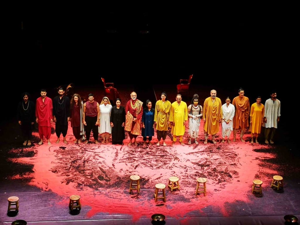 16 people in varied and brightly coloured Indian clothing, stand in a single row on a large stage. On the floor, they are standing across a large circle of red powder, spread out and marked by dancing feet. 