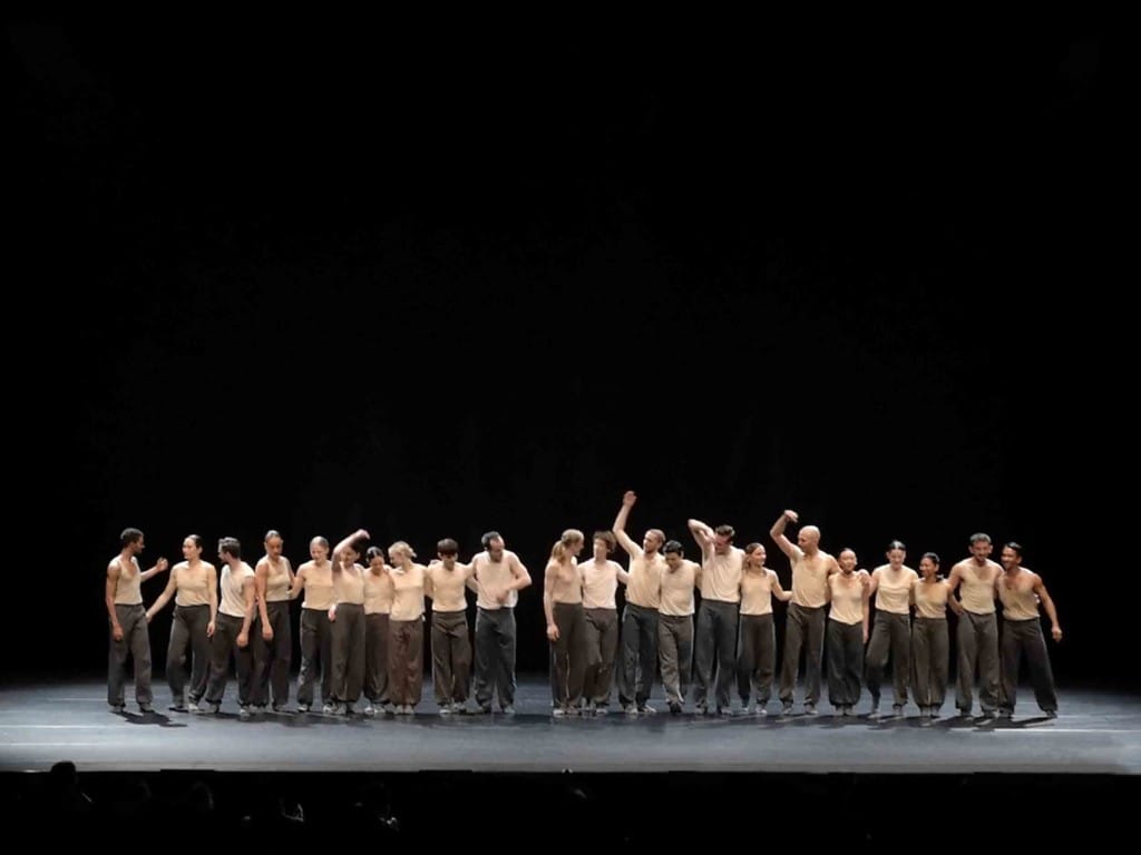 Around 20 dancers, each dressed in black trousers and cream singlet, stand in a line, many with their armes around each other, on the large blank stage at Sadler's Wells. 