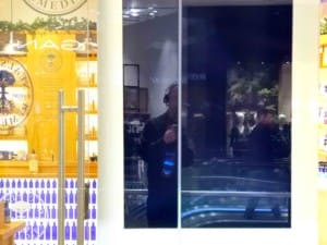 Reflected in a blank advertising screen we see a half cut-off image of a man wearing headphones and taking a photo of his reflection. Behind the screen is a brightly lit perfume shop.