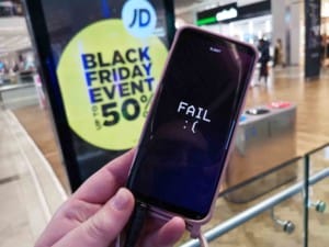 In the foreground, a mobile phone is held to show the screen contains the word FAIL. In the background is a digital display advertising a Blac Friday event sale of 50% off.