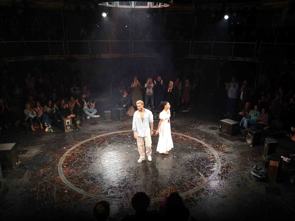 Two young people, dressed in white, stand slightly apart from each other on a circular stage. They are laughing, taking applause.