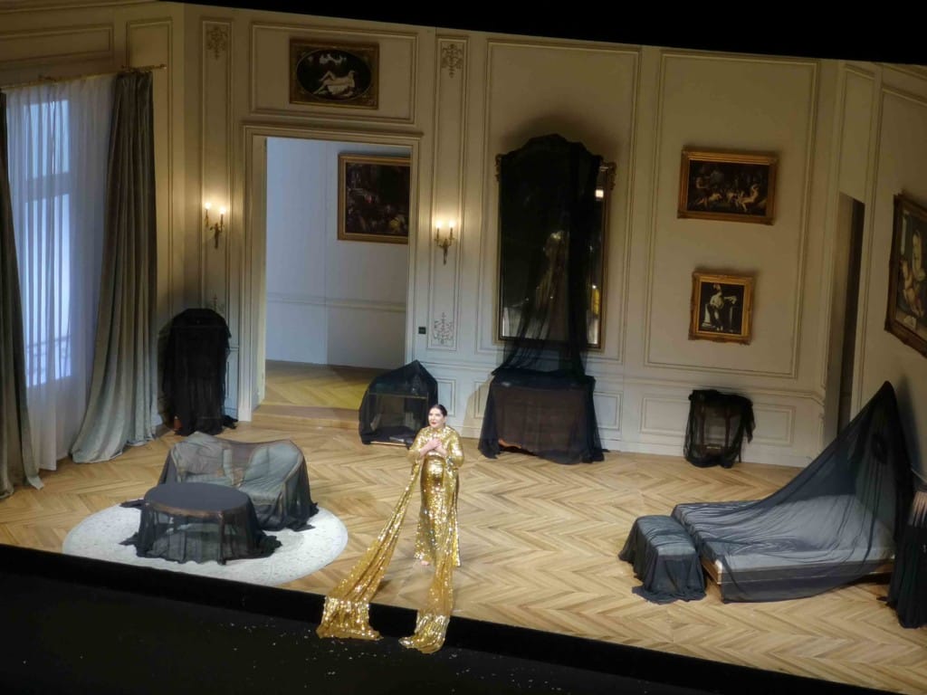 A stage set of a 19th century French hotel room, draped in black cloths as if in mourning. At the fore of the stage a lone female figure stands, dressed in a gold dress with oversized sleeves draped over the front of the stage.
