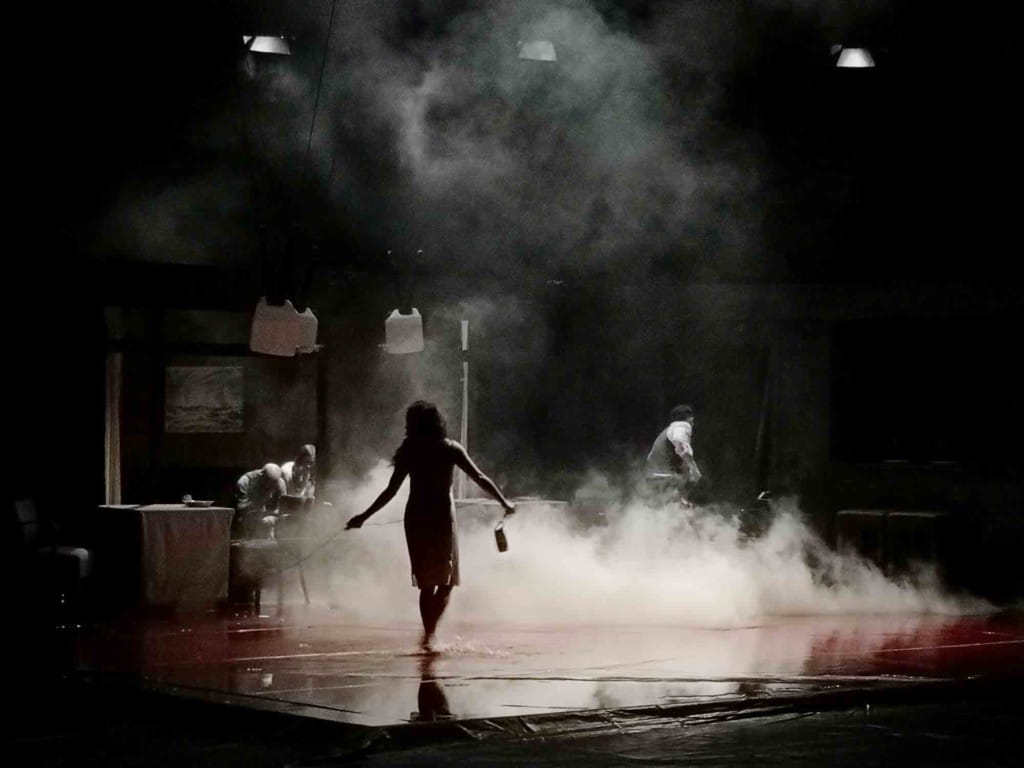 A lone, female figure, in silhouette, from behind. Barefoot she is walking through water on a stage. Steam appears to be rising from the stage in front of her.