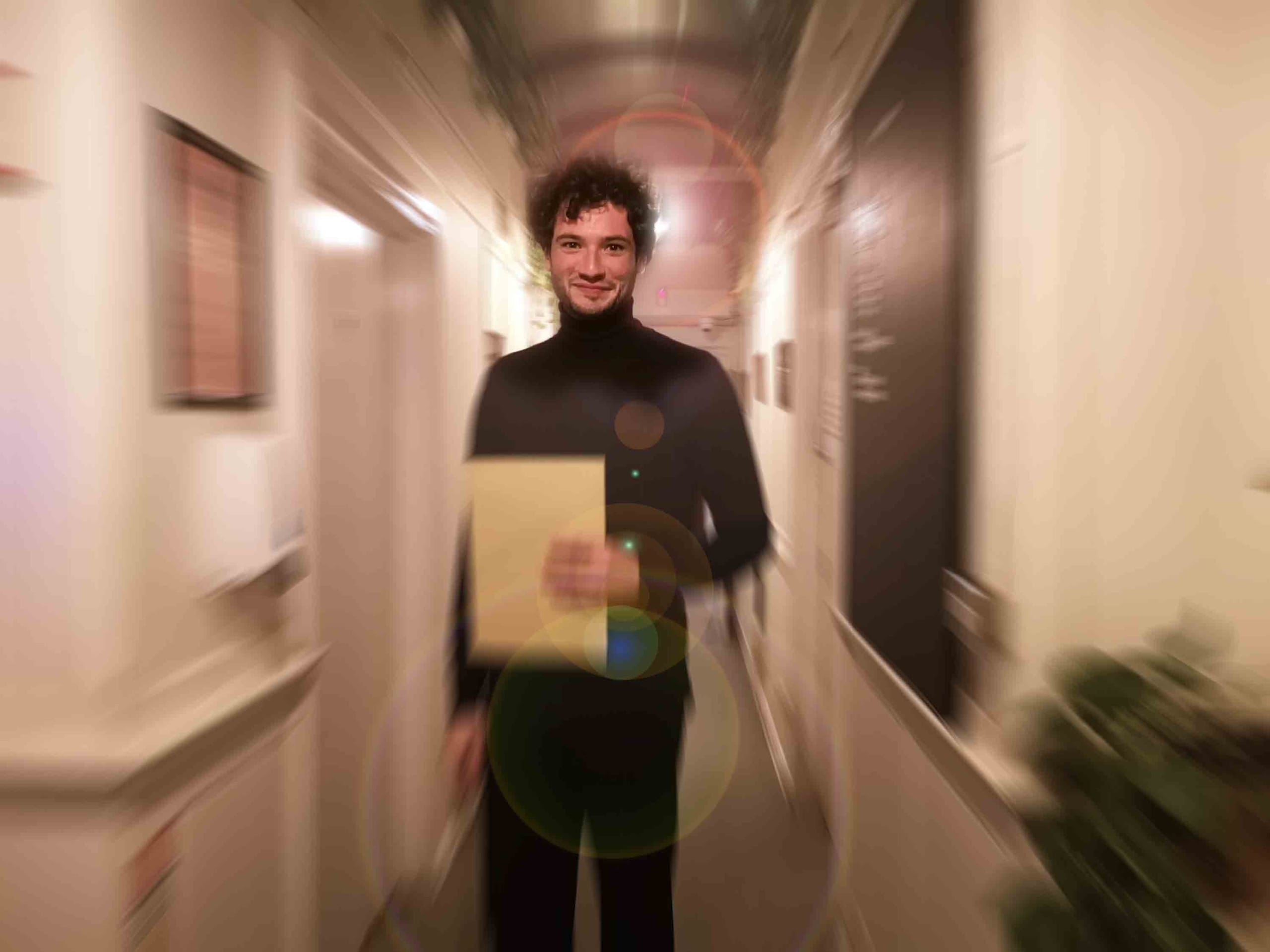 Jack Aldisert, a young white man, dressed in a black roll-neck, stands in a corridor holding a manila envelop. The image has been distorted so the corridor is blurred as if in motion.