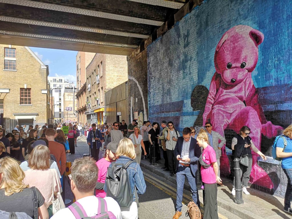 A busy section of street, partially under a railway bridge, in Shoreditch. A mural on the wall shows a large, beanie baby-esque bear, partially slumped over. The street is crowded/ Two big lines are formed, but there are also other people milling about in the middle. 