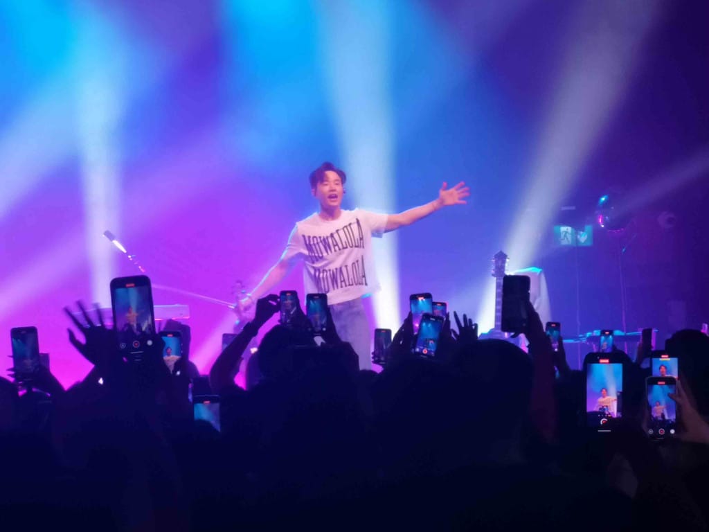Performer Henry Lau has arms outstretched in front of a crowd who all seem to have their illuminated phones held aloft with his image on it.