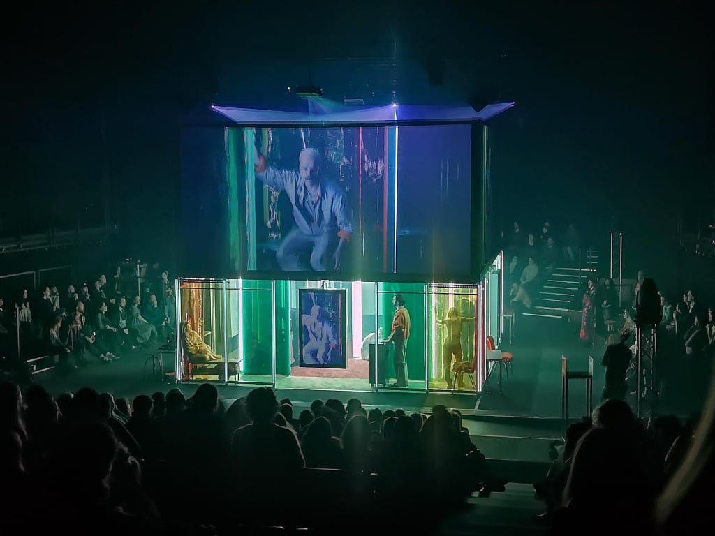 A brightly lit cube with actors at ground level and screens (featuring the actors) above. The cube is surrounded in all sides by a seated audience, in the dark.
