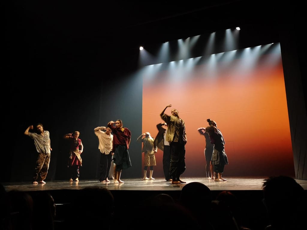 10 dancers, dressed in loose casual clothes hold a pose with right arm aloft and elbow bent. They stand in a grid formation on a lit stage.