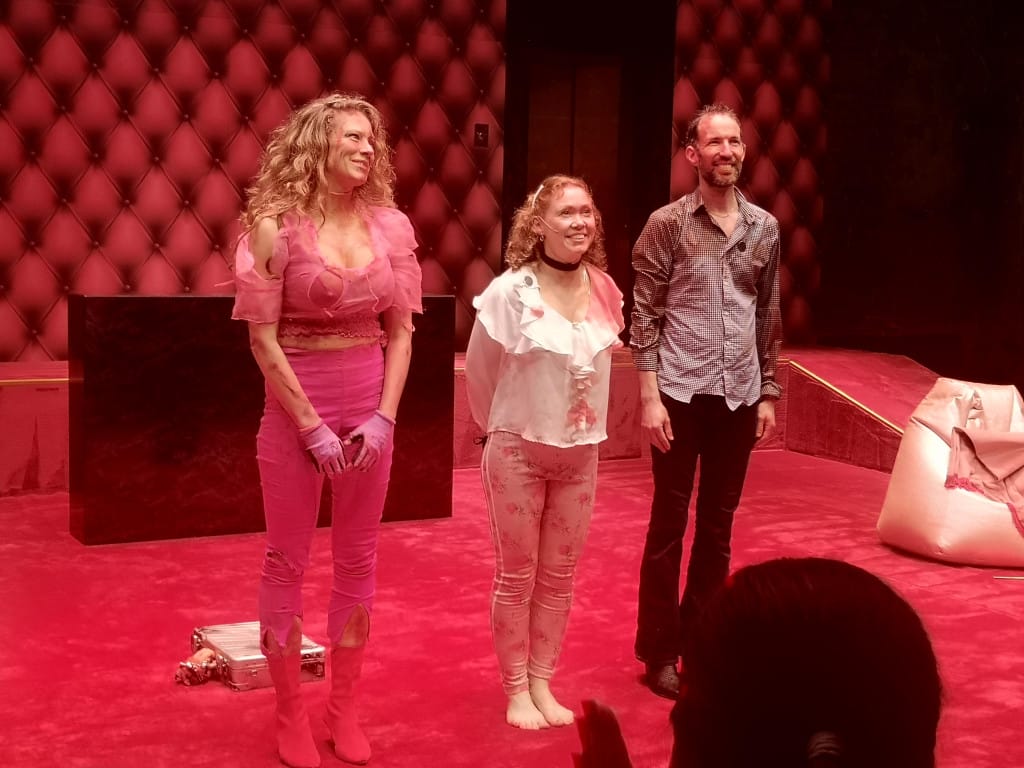 Three blood-soaked performers stand to receive applause on a pink carpeted stage.