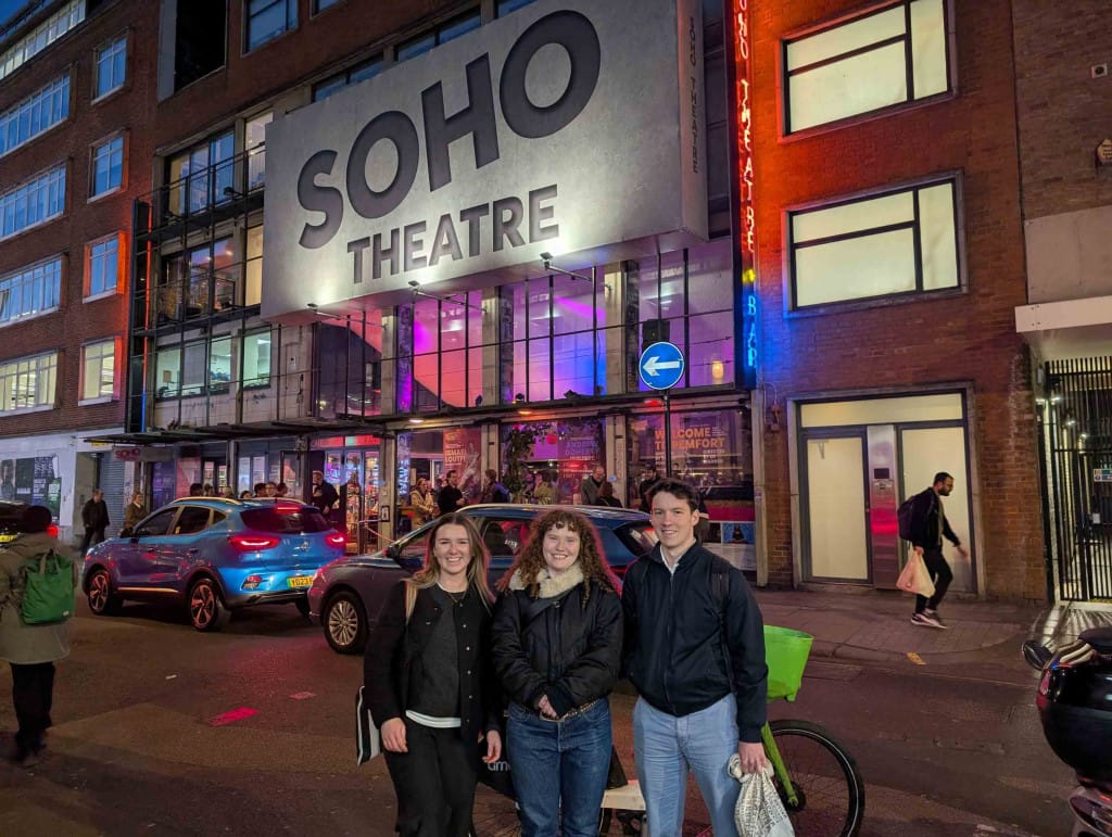 At night, three young people stand together, smiling. They are in a busy London Street in front of an illuminated Soho Theatre.