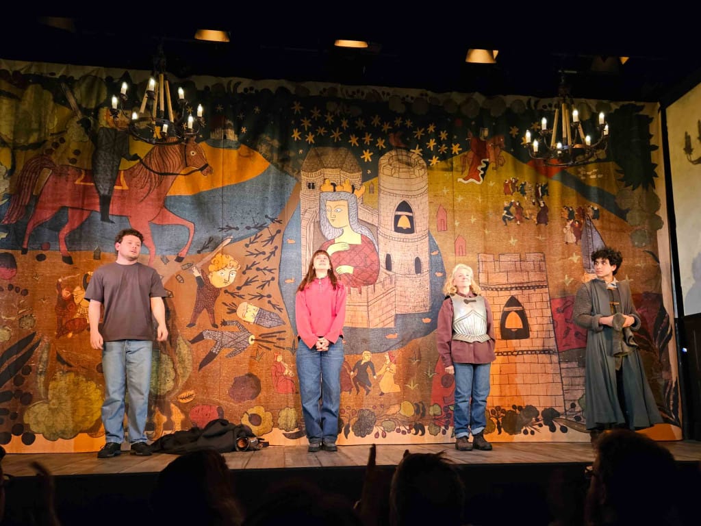 Four actors stand apart on stage to take their applause. Behind them is a dropped curtain that fills the space of the stage, on it is a slightly cartoon-like medieval illustration featuring a castle and a beheaded knight. 