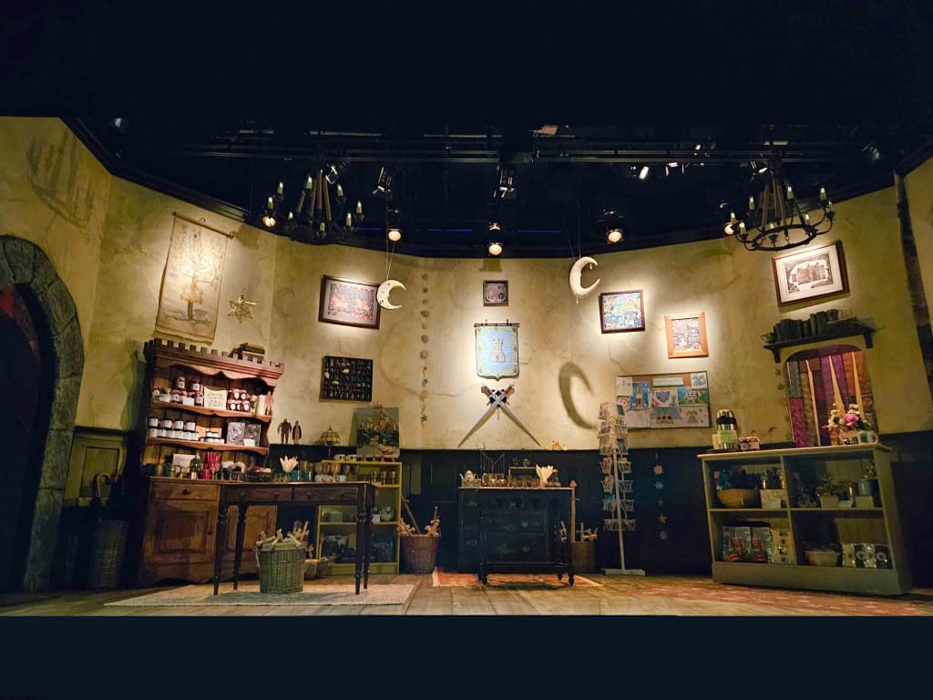 Theatre set that looks like the gift shop in a heritage attraction. The shelving is mismatched and there are medieval pictures and cross-swords on the back wall. 