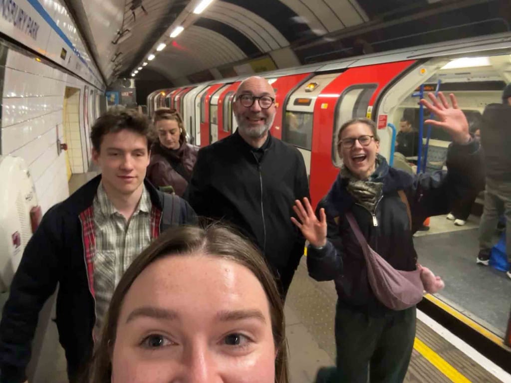 Beside a tube train, five people walk along the platform, smiling.