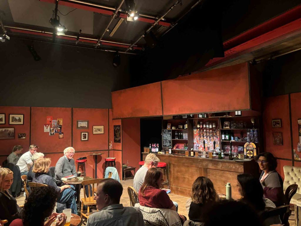 A theatrical set that looks like the bar of a British pub. There are people seated at tables and chairs in front of the bar.