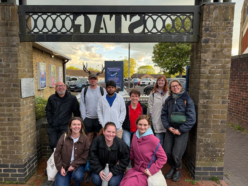 A group of nine smiling people posing for a photo together outdoors under a brick archway sign that reads 'STAG' in reverse.
