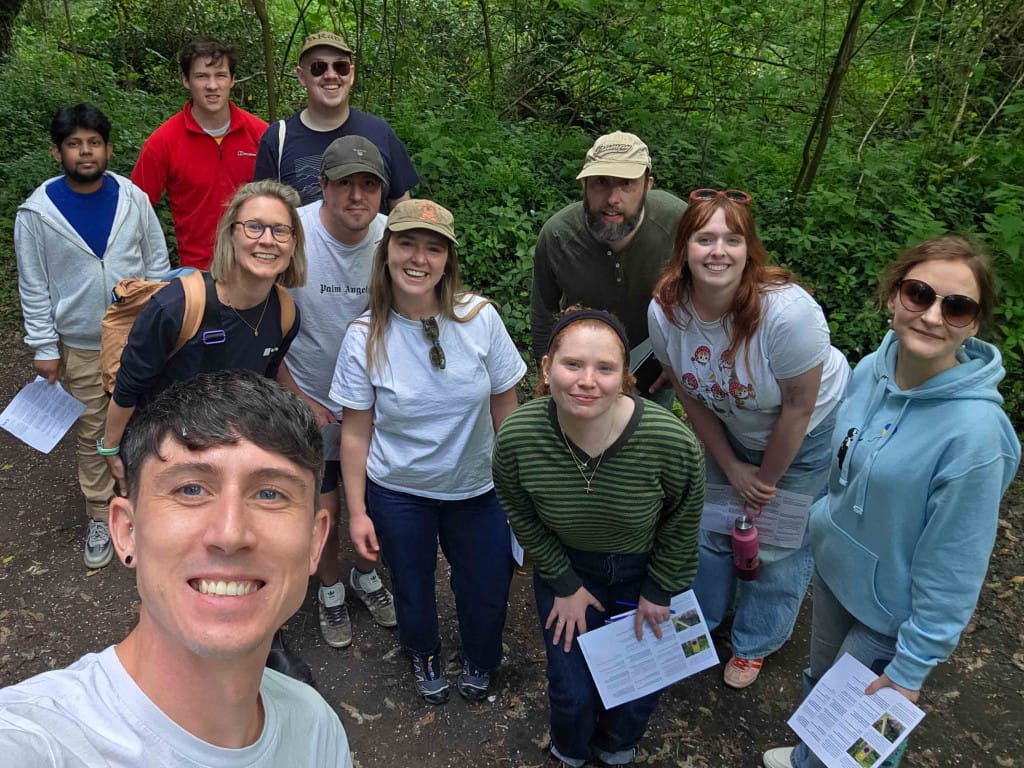 A man takes a selfie with a group of smiling, casually dressed people behind him. They are in a woodland setting and some are holding pieces of paper.