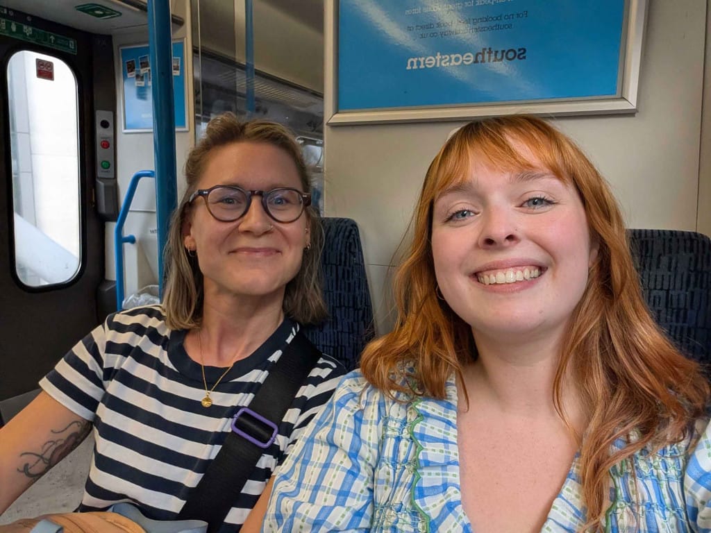 Two females, one wearing a stripy blue and white top and the other wearing a chequered blue and white dress, sat on a train.